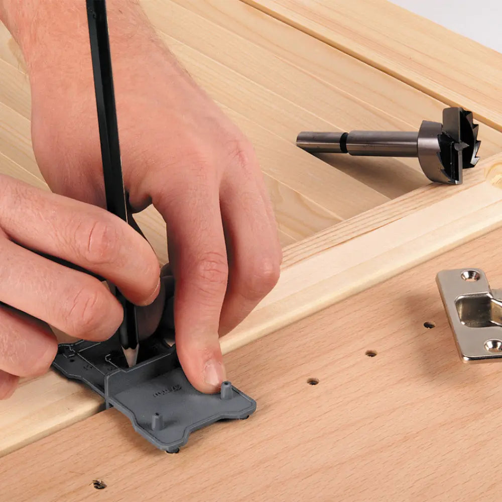 En primer plano, las manos de un carpintero marcando con un lápiz los agujeros de la plantilla antes de perforar con la broca